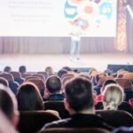 Audience attentively watching a presentation in a conference setting, with stage lights and speaker visible.